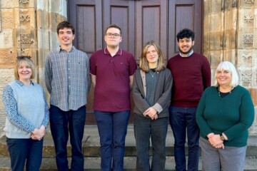Four students and two staff members pose on steps in front of an old door